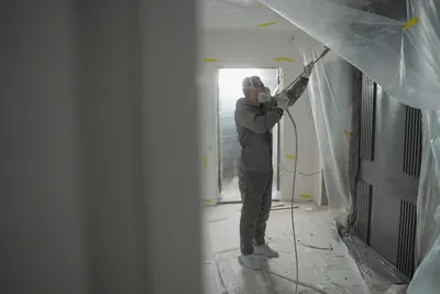 Worker in a gray suit and mask spraying inside a room sealed with plastic sheeting